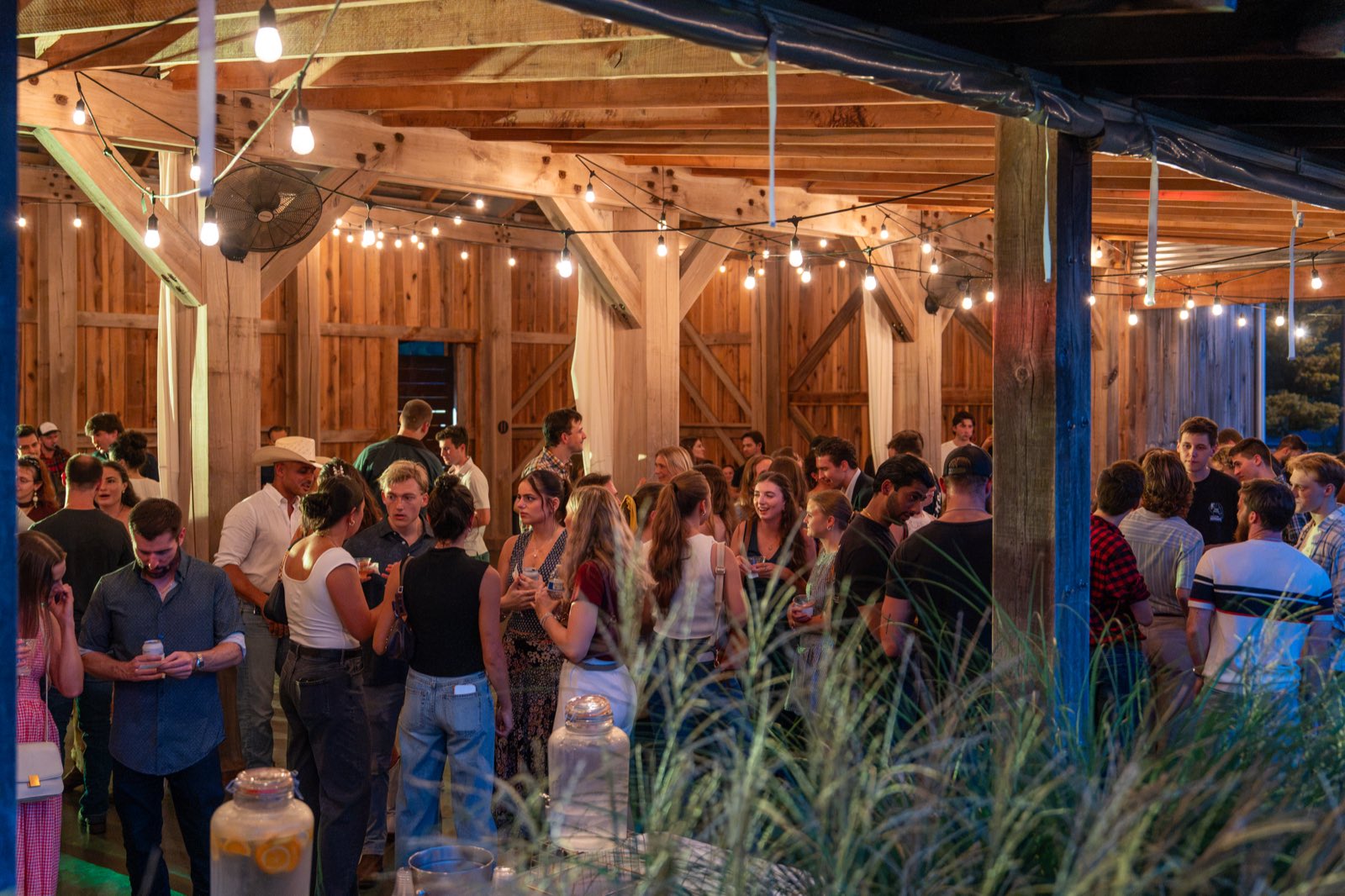 Packed night crowd talking under string lights inside the barn